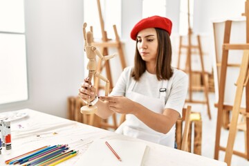 Young hispanic artist woman wearing french beret holding manikin at art studio.