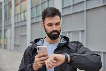 Serious bearded man checks time on smartwatch uses application to monitor fitness activity dressed in black anorak poses outdoors against blurred background. Peope sport and technology concept - obrazy, fototapety, plakaty