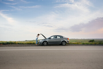 Fototapeta premium A young girl stands near a broken-down car in the middle of the highway during sunset and tries to call for help on the phone. Breakdown and repair of the car. Waiting for help.