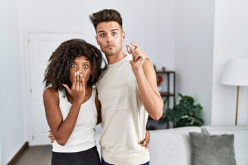 Young interracial couple holding keys of new home covering mouth with hand, shocked and afraid for mistake. surprised expression