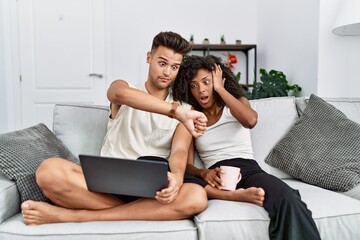 Young interracial couple using laptop at home sitting on the sofa looking at the watch time worried, afraid of getting late