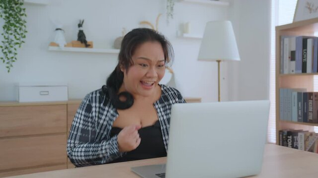 A big Asian woman sitting at her desk in front of a laptop computer celebrate offer sale good price. When she successfully offers a job to a client. Winner celebrate monetary victory concept.