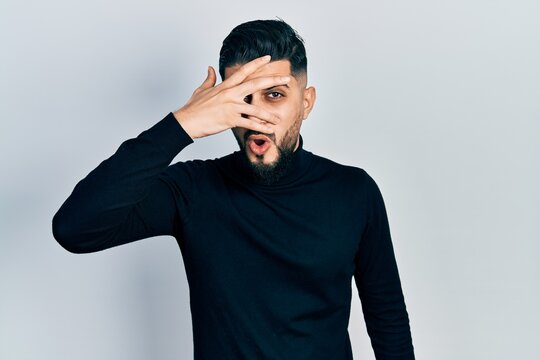 Handsome man with beard wearing turtleneck sweater peeking in shock covering face and eyes with hand, looking through fingers afraid
