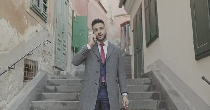 bearded young businessman with long coat and suitcase talking on the phone and going down the stairs in a medieval city