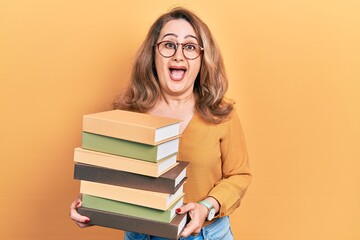 Middle age caucasian woman holding a pile of books celebrating crazy and amazed for success with open eyes screaming excited.