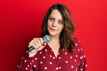 Young caucasian woman singing song using microphone thinking attitude and sober expression looking self confident