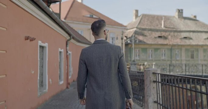 back view of elegant businessman with briefcase walking and looking to side outside in a medieval city 