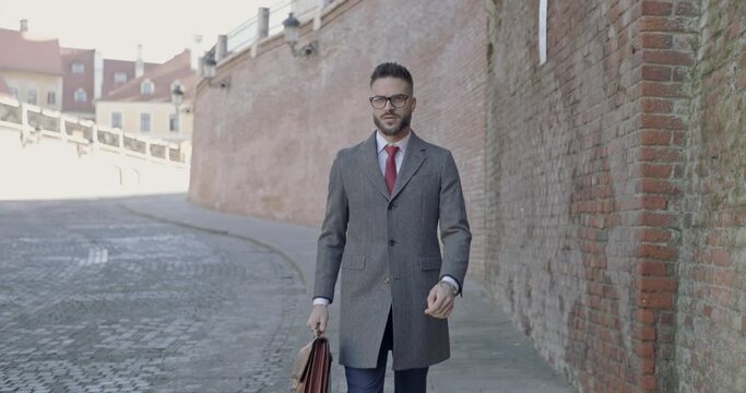 fashion model walking, looking away, holding a briefcase, arranging his glasses outdoor