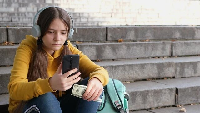 The teenager holds dollar bills and looks at the smartphone screen.