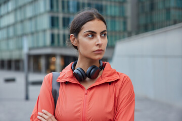 Photo of thoughtful woman wears windbreaker wears stereo headphones around neck returns fromm fitness training poses outdoors against blurred background. People and healthy lifestyle concept © WHstudio Leushin N