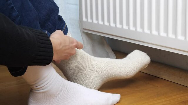 Woman pulling on woolen socks sitting near heating radiator at home. Expensive heating costs during cold season