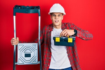 Young hispanic man holding tools box wearing hardhat by construction stairs clueless and confused...