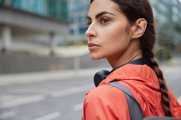 Cropped shot of thoughtful millennial girl with pigtail looks away pensively dressed in sportsclothes carried fitness mat necessary foor training going to cross road stands outdoors blurred background © WHstudio Leushin N