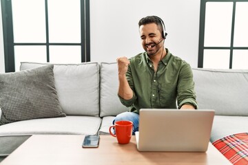 Young hispanic man with beard wearing call center agent headset working from home very happy and excited doing winner gesture with arms raised, smiling and screaming for success. celebration concept.