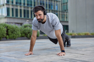 Fototapeta premium Outdoor shot of motivated strong bearded man does plank exercise demonstrates endurance poses against urban background listens music in headphones makes strength workout trains body does push ups