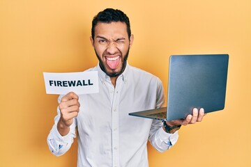 Hispanic man with beard holding computer laptop and firewall banner sticking tongue out happy with funny expression.