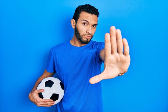 Hispanic man with beard holding soccer ball doing stop sing with palm of the hand. warning expression with negative and serious gesture on the face.