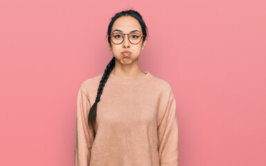 Young hispanic girl wearing casual clothes and glasses puffing cheeks with funny face. mouth...