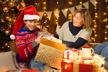 Fototapeta na wymiar Portrait of mother and son in new year decoration. Woman and teen boy opening the box, take out gifts, having fun and laughing. Holiday lights and christmas tree decorated with toys.