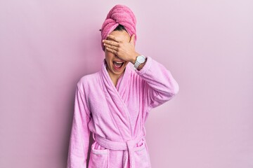 Young blonde woman wearing shower towel cap and bathrobe smiling and laughing with hand on face covering eyes for surprise. blind concept. - obrazy, fototapety, plakaty