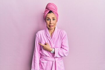 Fototapeta premium Young blonde woman wearing shower towel cap and bathrobe pointing aside worried and nervous with forefinger, concerned and surprised expression