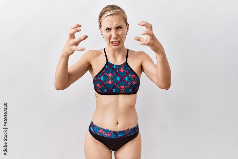 Sticker Young blonde woman wearing sporty bikini over isolated background shouting frustrated with rage, hands trying to strangle, yelling mad