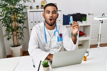 Young indian doctor holding medical asthma inhaler at the clinic scared and amazed with open mouth for surprise, disbelief face - obrazy, fototapety, plakaty