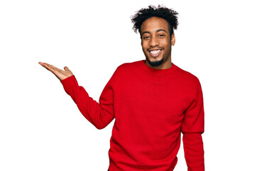 Young african american man with beard wearing casual winter sweater smiling cheerful presenting and pointing with palm of hand looking at the camera.