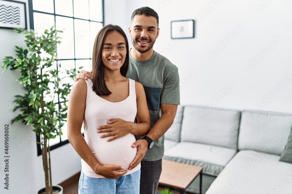 Poster young interracial couple expecting a baby, touching pregnant belly with a happy and cool smile on fa