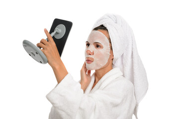 Caucasian middle aged woman with wet sheet mask on her face looks in the mirror, she is dressed in a white bathrobe with a towel on her head, white isolated background.