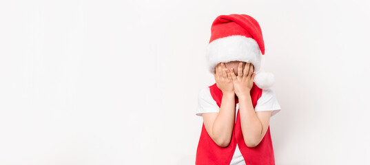 Portrait of crying little child in red Santa Claus hat isolated on white background. European 5 year boy. Without gifts. Face is covered with hands. Bad mood. Christmas. Close-up. Sleepy or tired kid