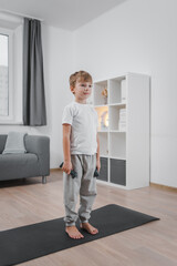 Sports hall at home. Portrait of Caucasian child boy doing fitness with dumbbells in living room. The child is dressed in a white t-shirt and gray pants.