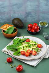 Fresh salad with avocado, cherry tomatoes, arugula, mizuna, chard and pumpkin seeds on a plate on a green background. Vegetarian vitamin food. Vertical view