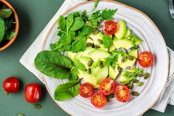 Fresh salad with avocado, cherry tomatoes, arugula, mizuna, chard and pumpkin seeds on a plate on a green background. Vegetarian vitamin food. Top view. Close-up