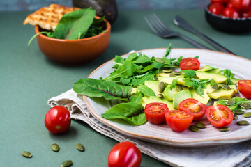 Ready-to-eat salad with avocado, tomatoes, arugula leaves, mizuna, chard and pumpkin seeds on a plate  on a green table. Vegetarian vitamin food. Copy space