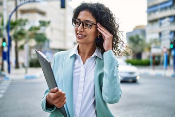 Fototapeta premium Young hispanic business woman wearing professional look smiling confident at the city holding worker clipboard