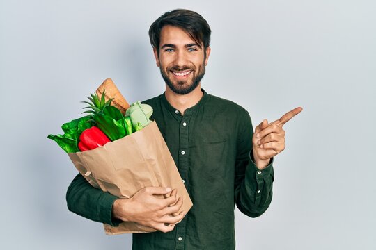 Young hispanic man holding paper bag with bread and groceries smiling happy pointing with hand and finger to the side
