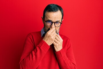 Middle age hispanic man wearing casual clothes and glasses smelling something stinky and...