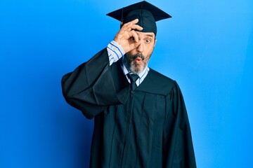 Middle age hispanic man wearing graduation cap and ceremony robe doing ok gesture shocked with surprised face, eye looking through fingers. unbelieving expression.