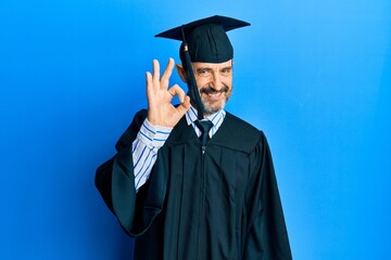 Middle age hispanic man wearing graduation cap and ceremony robe smiling positive doing ok sign with hand and fingers. successful expression.