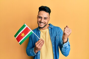 Young arab man holding suriname flag screaming proud, celebrating victory and success very excited...