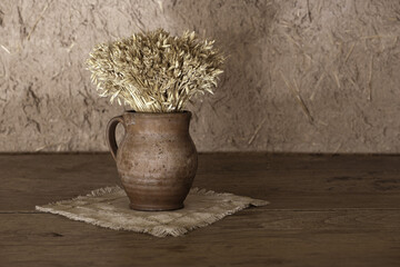 Abstract rural composition of clay vase with a bouquet of dried cereals standing on gray linen tablecloth and old, grunge and aged wooden desk in front of clay wall with copy space. HDR image