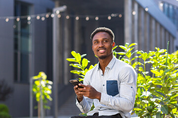 Cheerful african american guy using mobile phone sitting on bench in city park outdoors. Happy black man in casual clothes with a smartphone in hands chat on urban modern background on street outside
