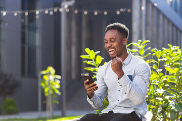 happy african american man in casual clothes sitting on bench outdoors with mobile phone in hand...