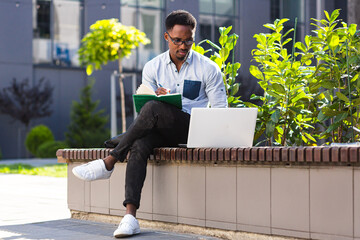 Young african american male student sitting in a city park on a bench with laptop and notebook...