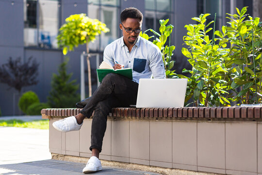 Young african american male student sitting in a city park on a bench with laptop and notebook studying online outdoors Man freelancer in glasses with books learn working remotely in street e-learning