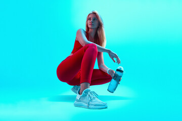 Young beautiful blonde sports girl in red sportswear squatting on a bright neon blue background...