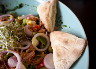 tuna salad with onion, cucumber, tomatoes, sweet red pepper, cucumbers and micrograins with honey sauce and two triangular loaves lies in a turquoise plate on a black background