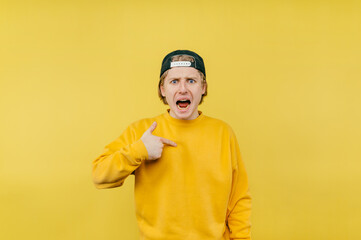 Dissatisfied young man points a finger at himself and looks at the camera with an evil face, isolated on a yellow background.