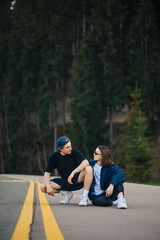 Stylish couple of hikers man and woman sitting on the asphalt in the mountains on a background of coniferous trees and talking. Vertical.